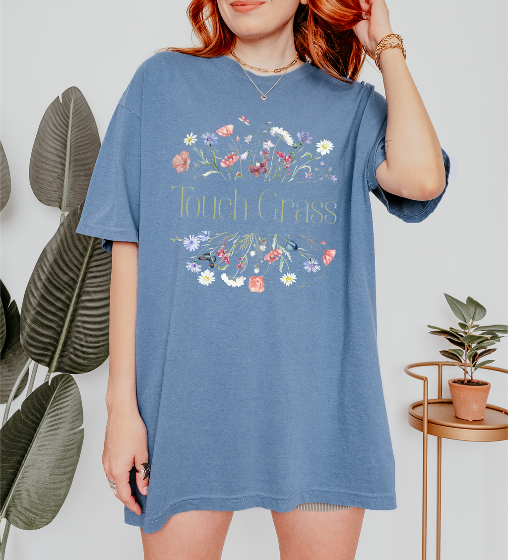 Person wearing a blue t-shirt with floral design and text that says touch grass, standing indoors with plants in the background.