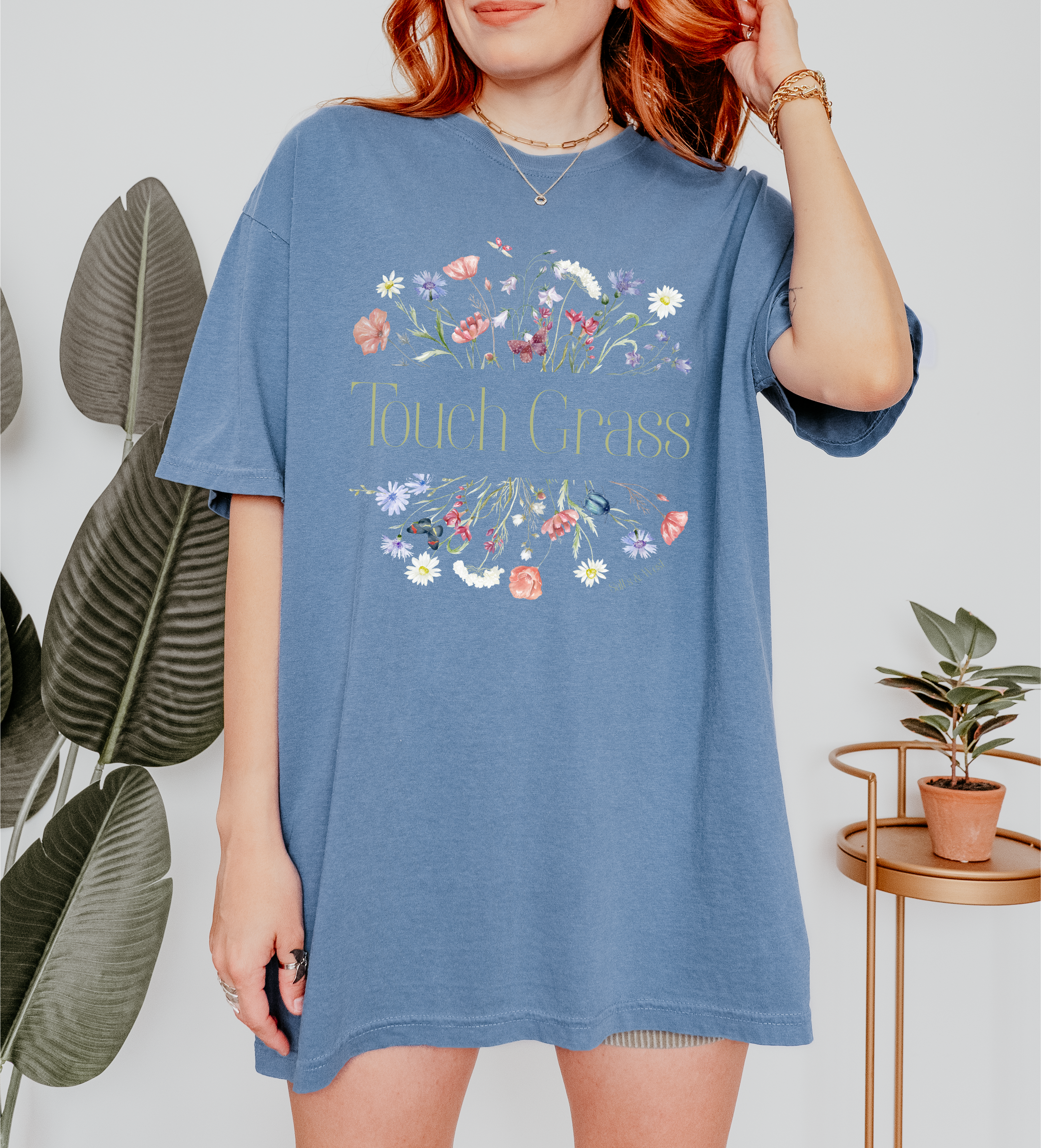 Person wearing a blue t-shirt with floral design and text that says touch grass, standing indoors with plants in the background.